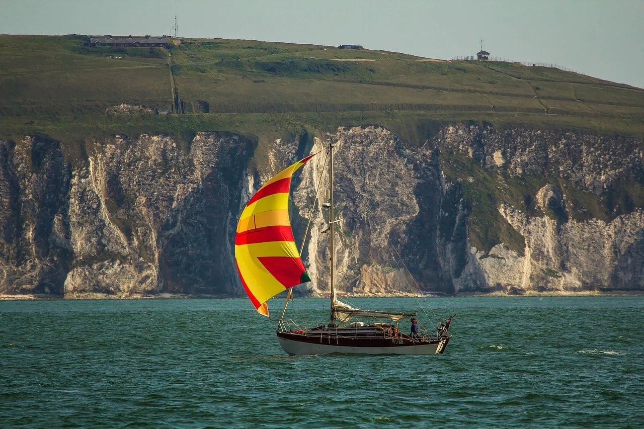 A small cruising yacht under a red and yellow striped spinnaker, sailing past white chalk cliffs topped with green grass