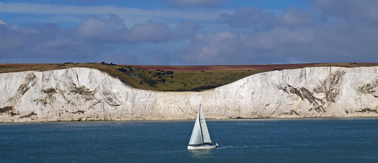 A small white-sailed yacht sailing past the white chalk cliffs of Dover on a blue sea under a cloudy sky